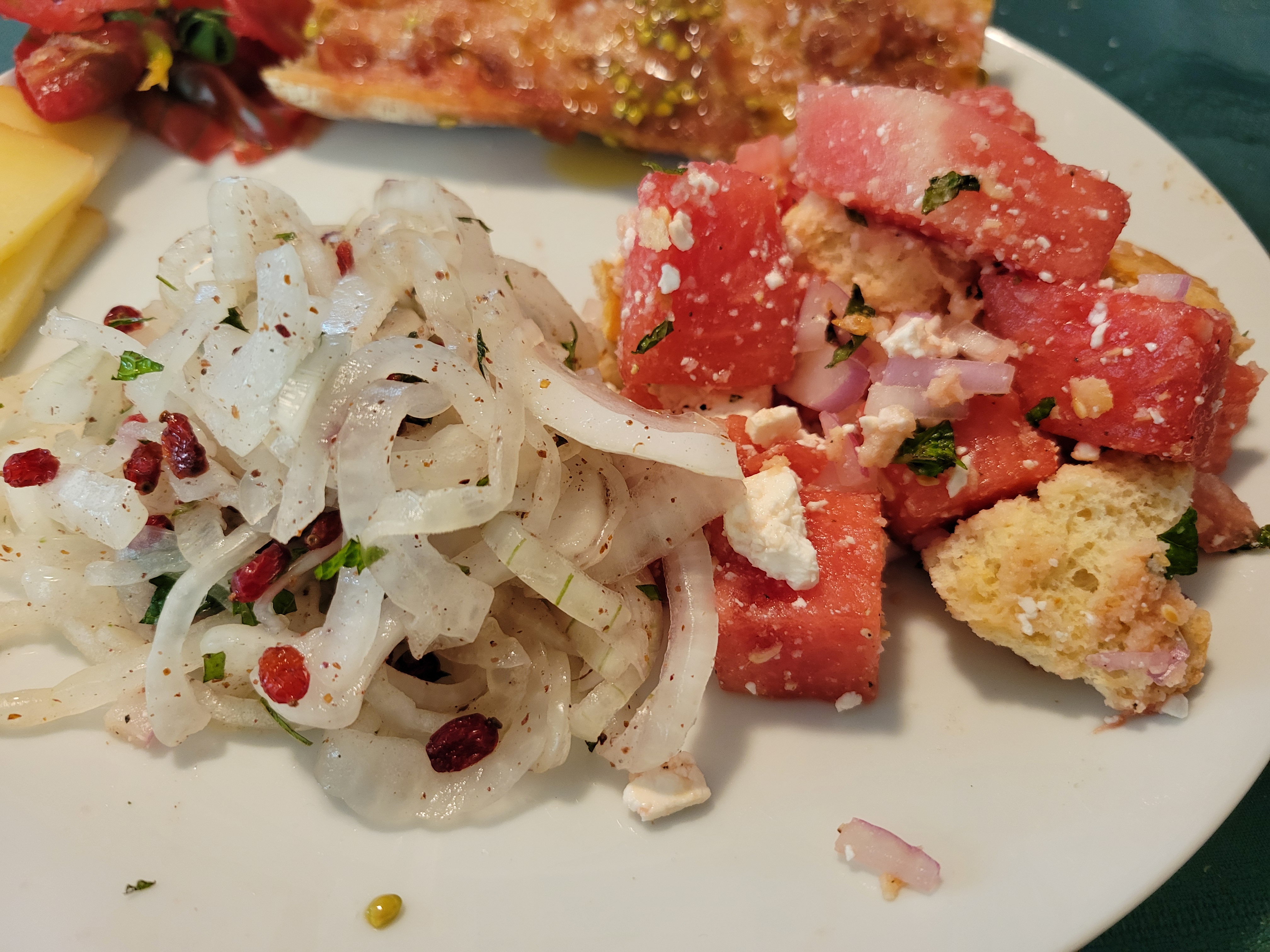 an onion salad and a watermelon salad on a plate, with tomato bread and cheese in the background